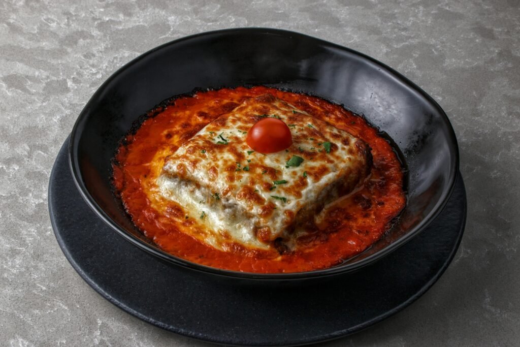 Ultimate Baked Lasagna Tasty baked lasagna with cherry tomato and cheese, served in a black bowl with rich tomato sauce.